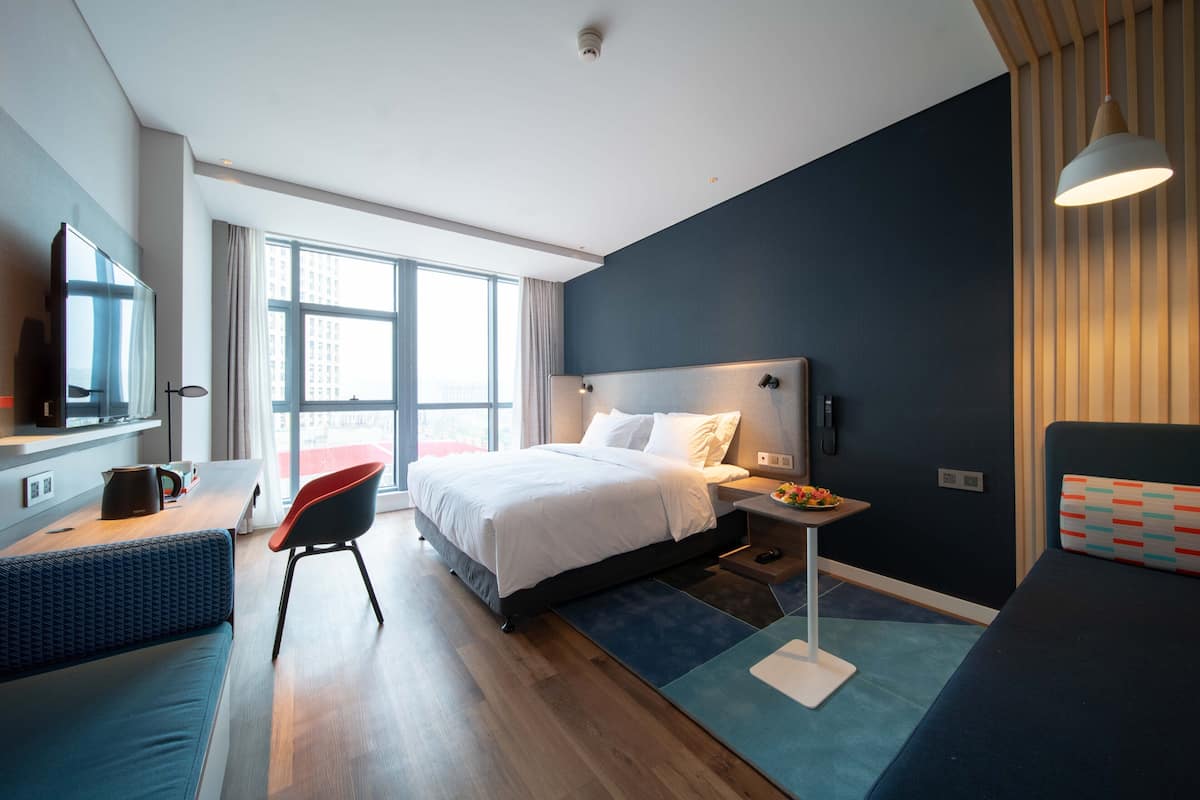 A modern hotel room with a large bed, a desk with a chair, a TV, and a window with curtains.