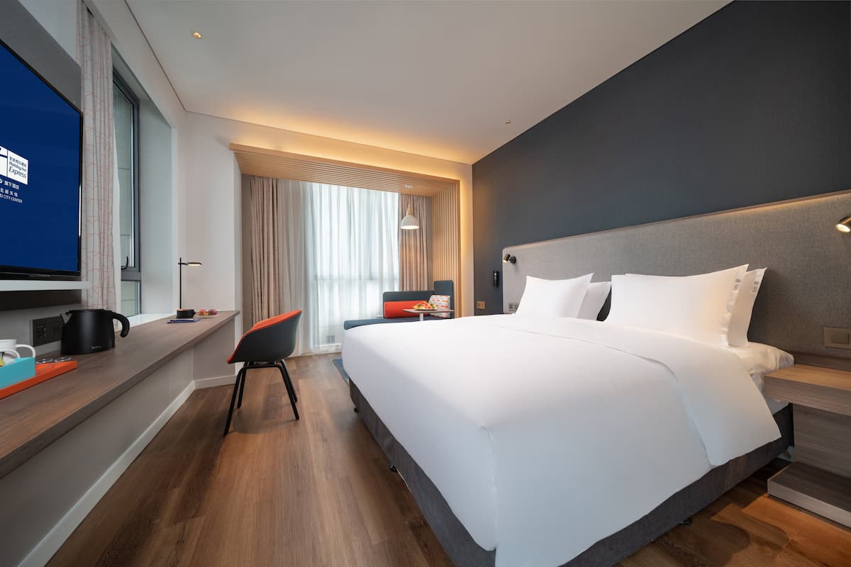 A modern hotel room with a large bed, a TV, a desk with a kettle, and a view of the city center.