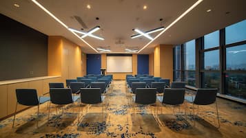 Una sala de conferències amb files de cadires, una pantalla de projector i un gran finestral amb vista a la ciutat.
