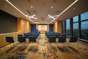 Un salón de conferencias con filas de sillas, una pantalla de proyector y un amplio ventanal con vistas a la ciudad.