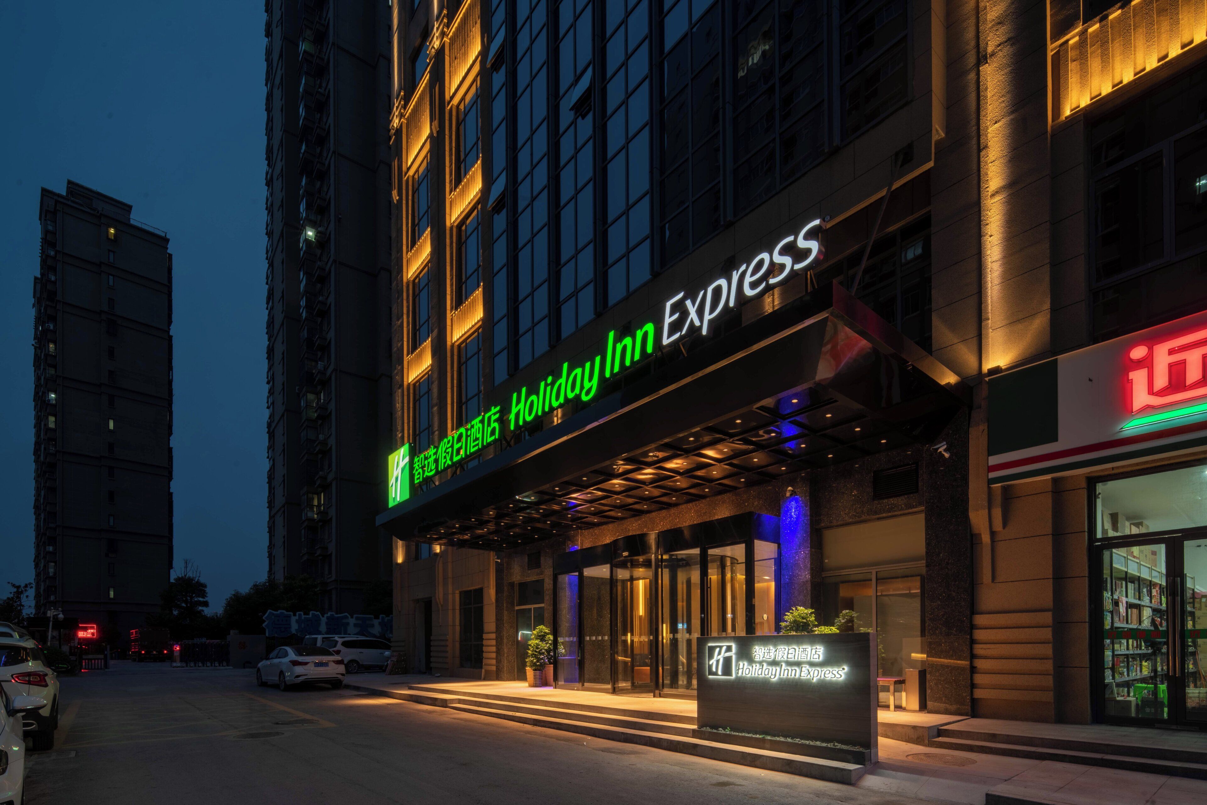 A hotel entrance with a sign for Holiday Inn Express, a glass door entrance, and a parking area with cars.