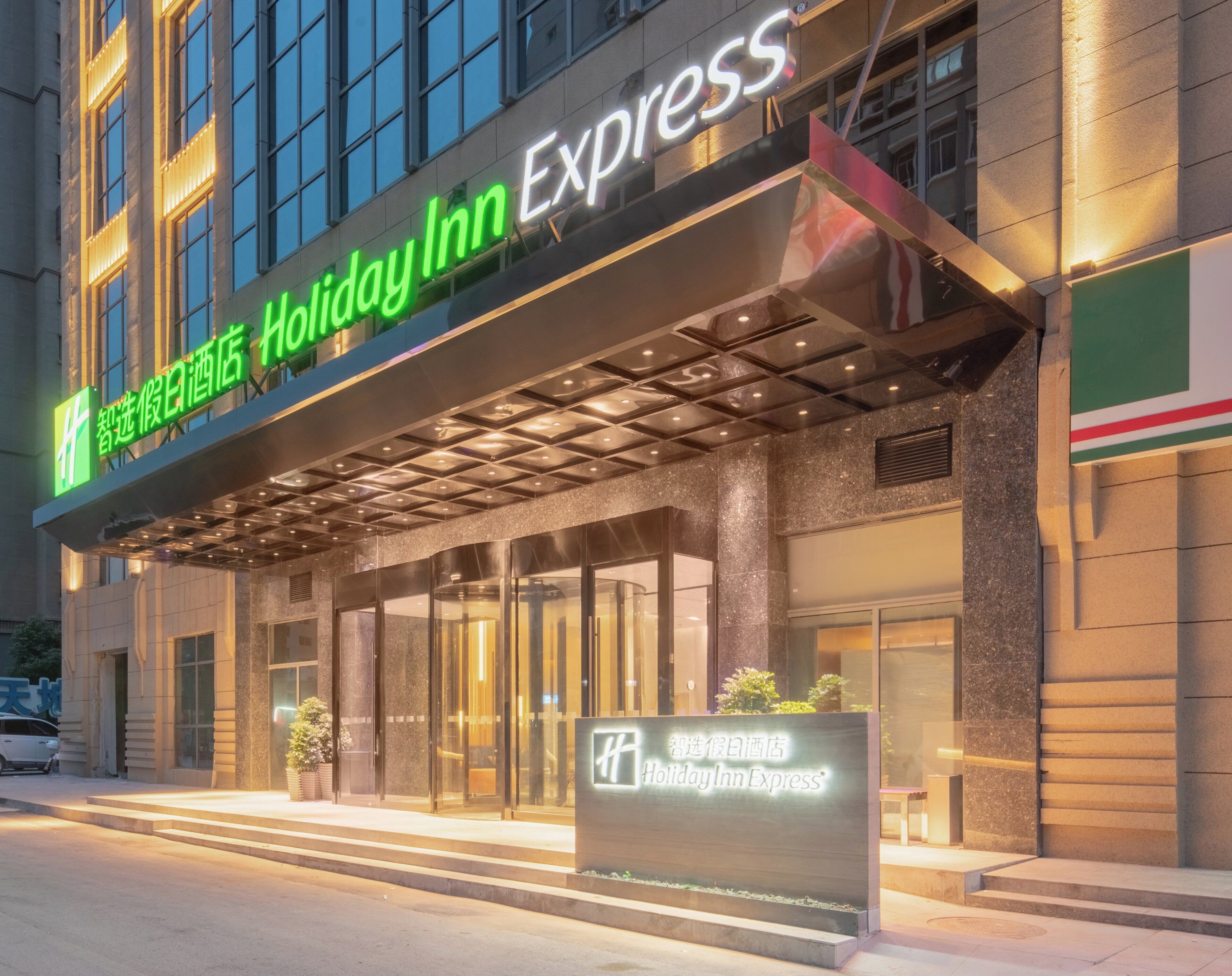 Entrada de un hotel con un llamativo letrero de 'Holiday Inn Express', una puerta de vidrio y un pequeño cartel con el nombre del hotel en inglés y chino.