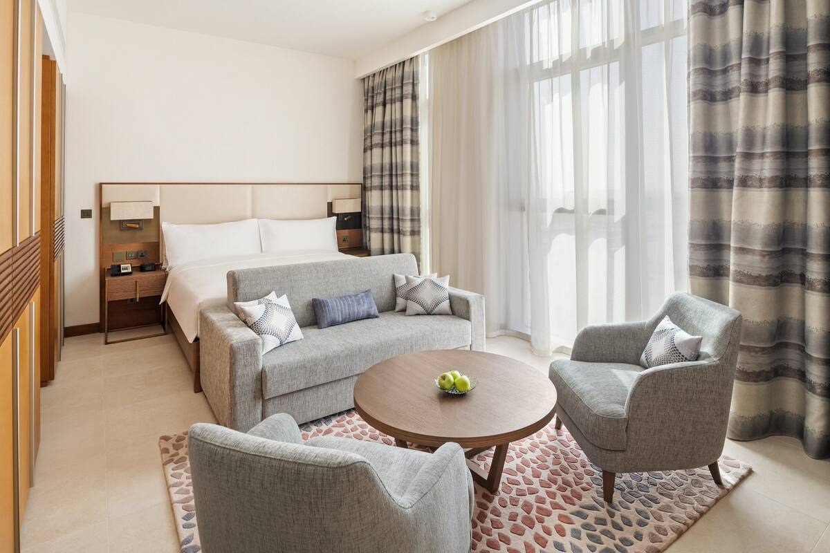 Une chambre d’hôtel moderne, équipée d’un lit, d’un canapé, de deux fauteuils, d’une table basse et d’une petite table d’appoint.