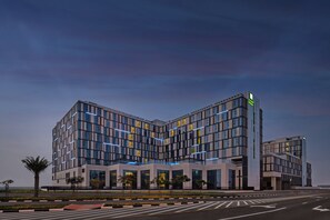 A modern hotel with a corner building design, featuring large windows and a prominent entrance. - Staybridge Suites Dubai Al-Maktoum Airport by IHG (Dubai)