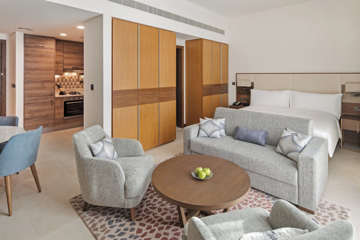 Ein Hotelzimmer mit einem Bett, zwei Sesseln, einem Couchtisch mit Äpfeln, einer Essecke mit Tisch und Stühlen sowie einer Küchenzeile.