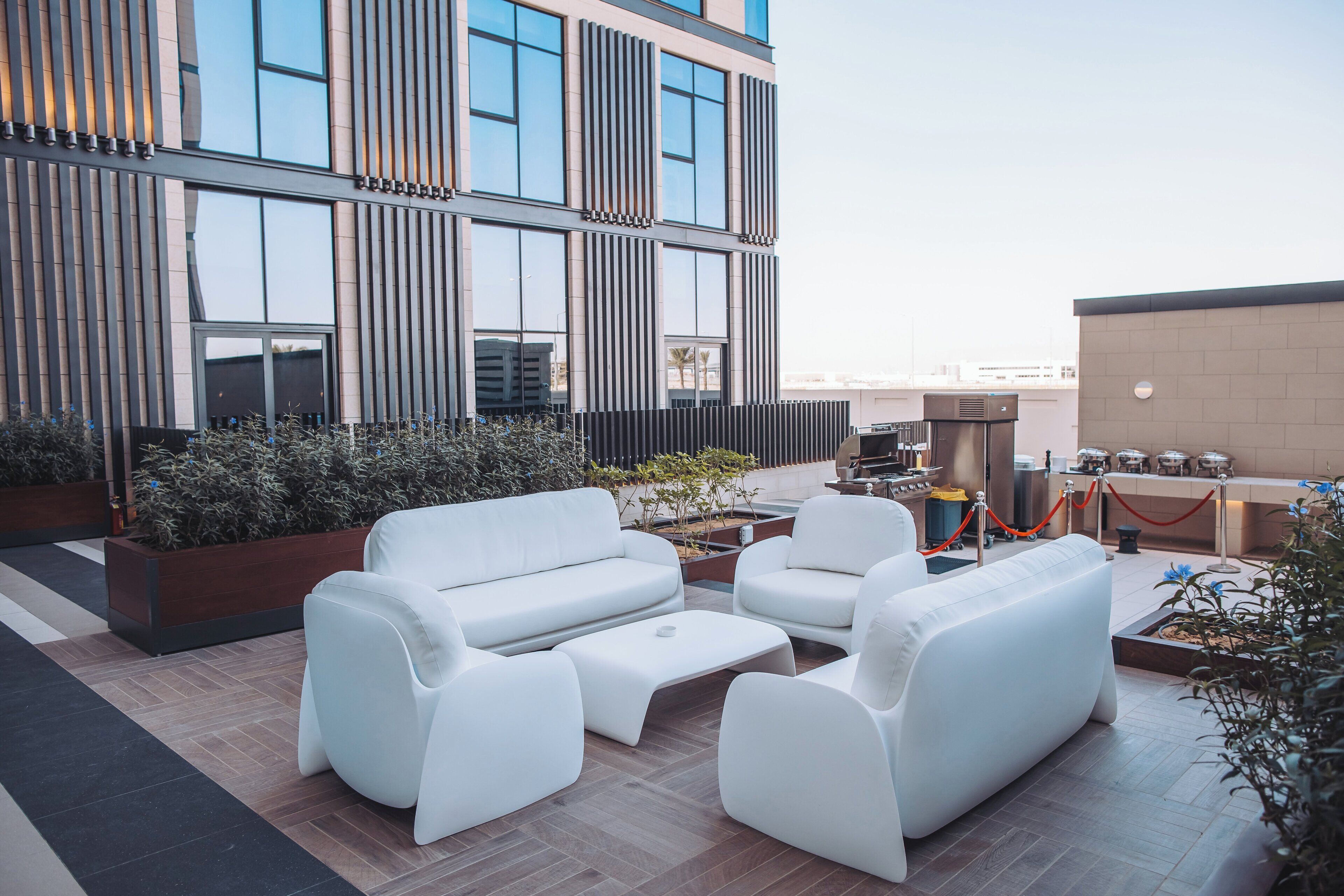 A rooftop lounge area with white sofas and a coffee table, a small table with a laptop, and a buffet setup.