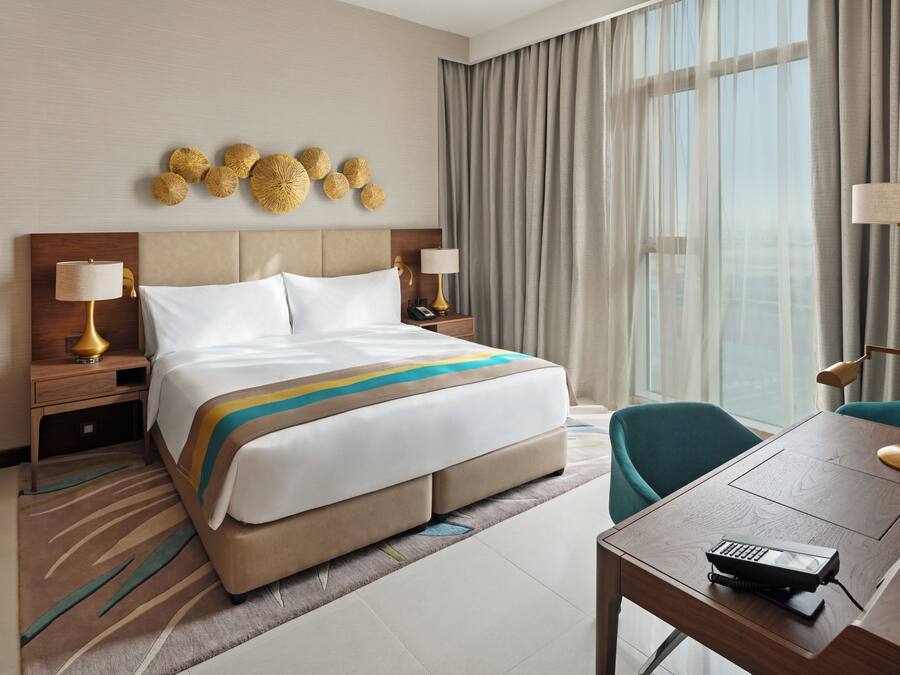 A hotel room with a large bed, two bedside tables with lamps, a desk with a telephone, and a view of the outdoors through the window.