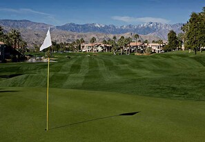 A golf course with a flagstick, well-manicured grass, and a backdrop of mountains.