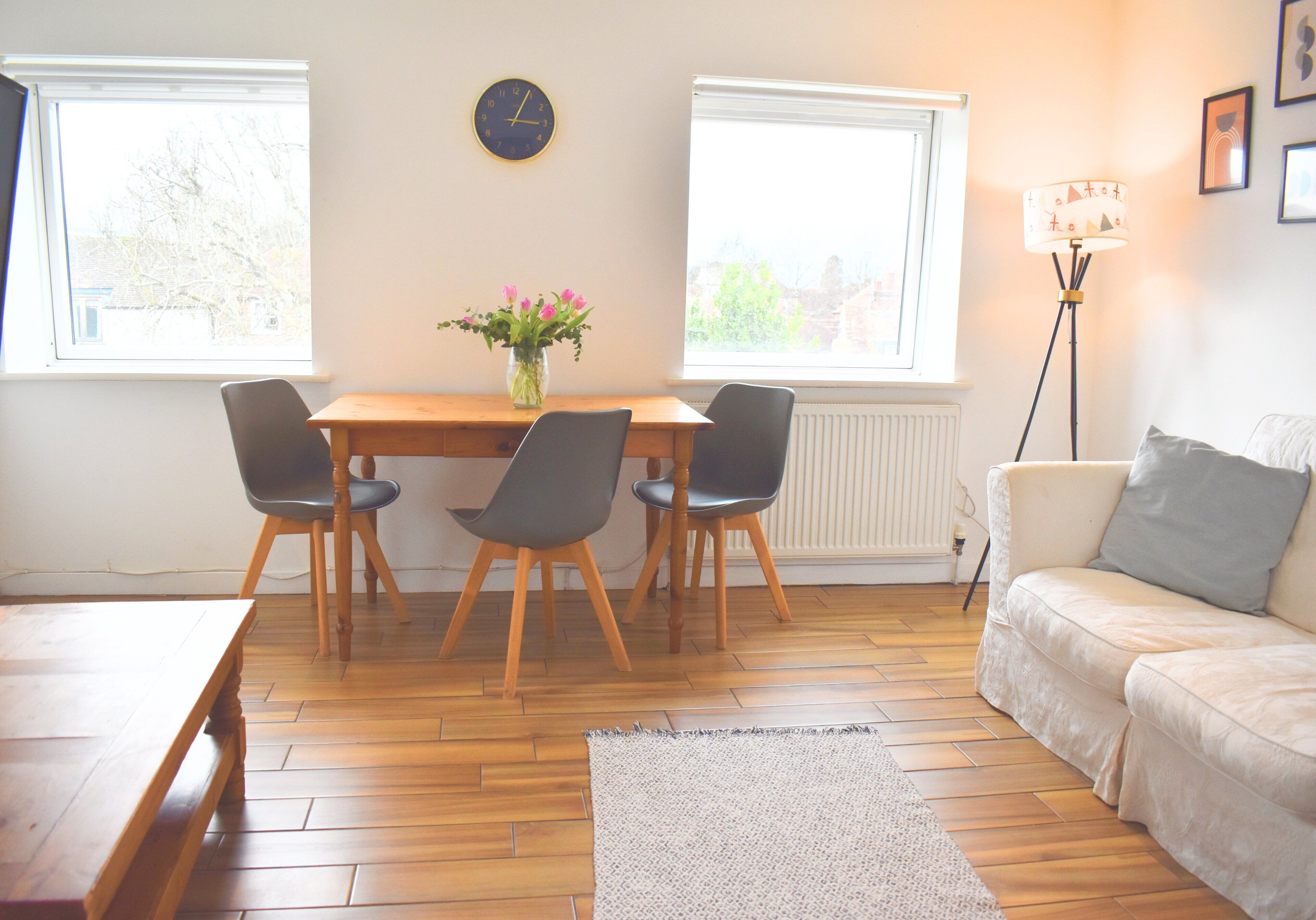 Una habitación con una mesa de comedor de madera, dos sillas, un sofá, una lámpara de pie y un reloj en la pared.