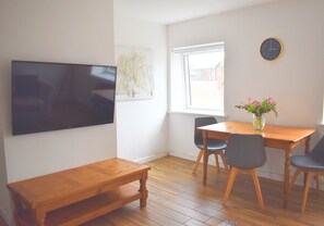 Une pièce comprenant une table en bois, des chaises, un banc, un téléviseur à écran plat, une fenêtre et une horloge murale.