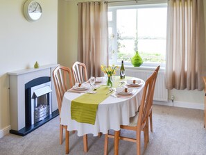 Een eetkamer met een ronde tafel gedekt voor vier personen, een open haard en uitzicht op de omgeving door grote ramen.