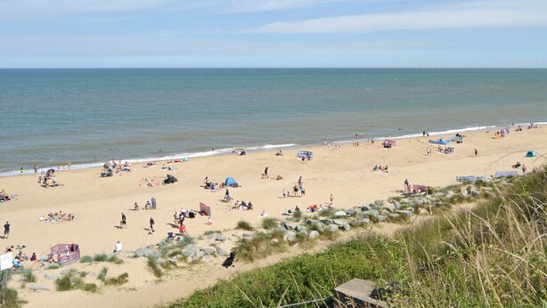 Ein Strand mit badenden Personen, Sonnenschirmen, Zelten und einem felsigen Küstenabschnitt mit Vegetation.