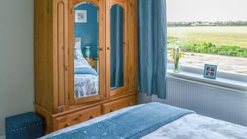 Een slaapkamer met een houten garderobe, een bed met bloemenbeddengoed, een raam met uitzicht op een veld, en een klein fotolijstje op de vensterbank.