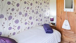 Ein Schlafzimmer mit floralem Wandpapier, einem Bett mit einem violetten Handtuch, einem Holzschrank und einem Nachttisch mit Lampe und einer Uhr.