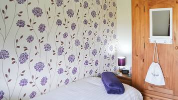 A bedroom with a floral wallpaper, a bed with a purple towel, a wooden wardrobe, and a bedside table with a lamp and a clock.
