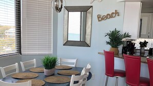 Un comedor con una mesa dispuesta para seis personas, rodeada de sillas blancas. Hay una zona de cocina con taburetes de bar rojos y una encimera con electrodomésticos.