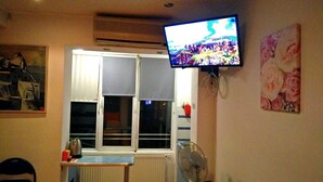 Una habitación con televisión montada en la pared, una ventana con persianas, un radiador y un ventilador.