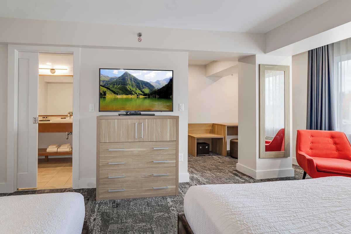 A hotel room with two beds, a TV mounted on the wall, a desk, a chair, and a bathroom visible through an open door.