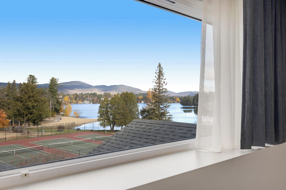 A view from a window overlooking a tennis court with a lake and mountains in the background.