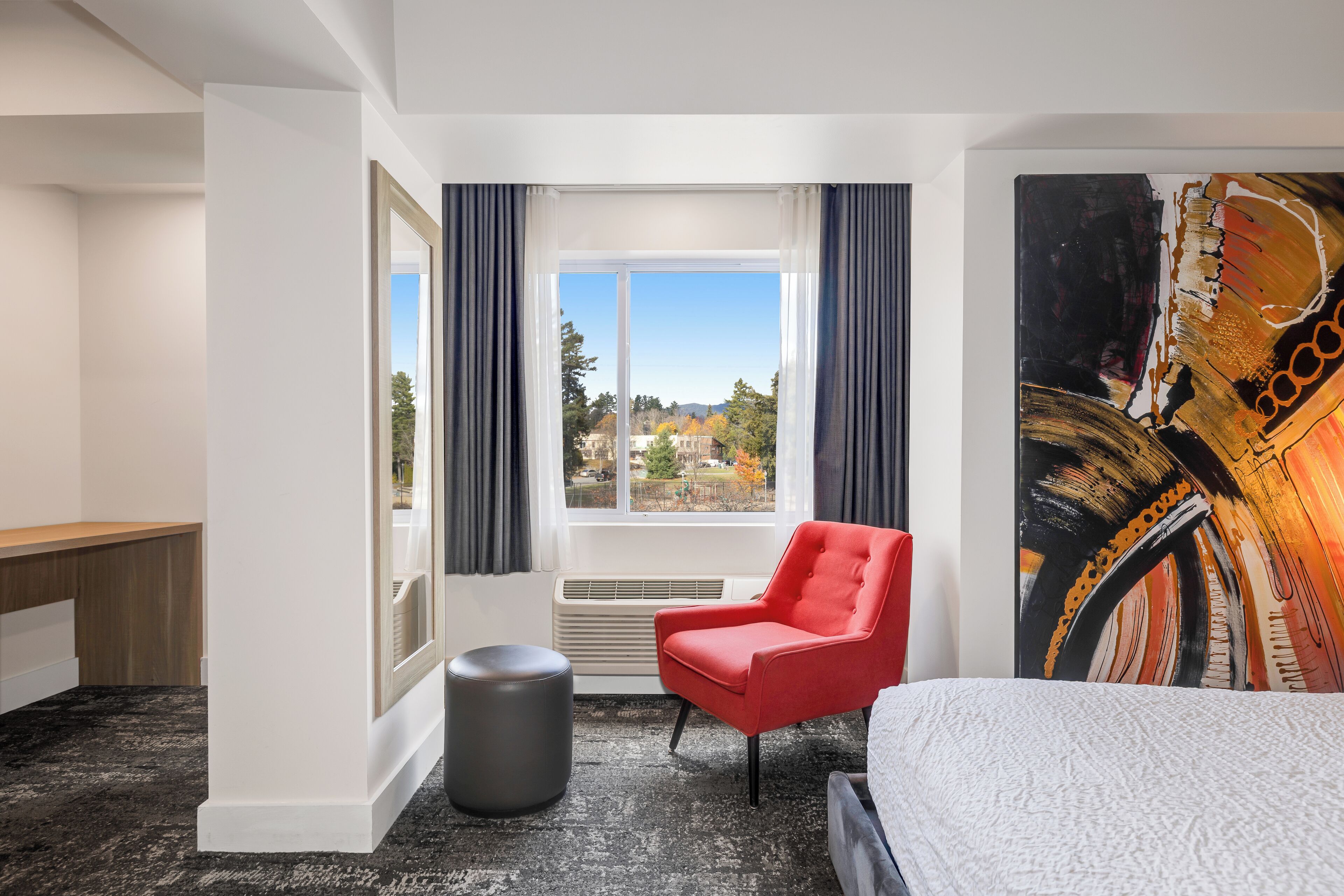 A hotel room with a bed, a red armchair, a large window with curtains, a painting on the wall, and a small round stool.