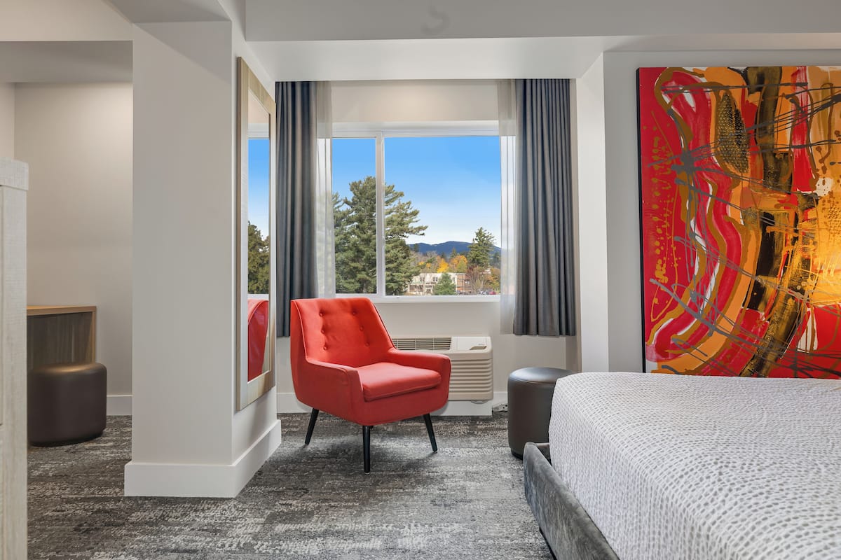 A hotel room with a large bed, a red armchair, a window with a view of trees and mountains, and a colorful abstract painting on the wall.