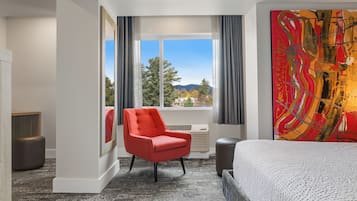 Un dormitorio de hotel con una cama grande, un sillón rojo, un ventanal con vistas a árboles y montañas, y un cuadro abstracto de colores en la pared.