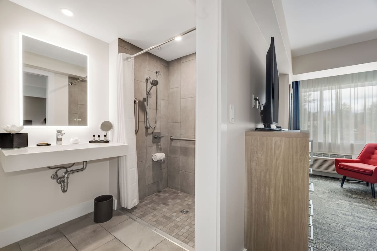 A modern bathroom with a large mirror, shower, and a red chair in the adjacent living area.