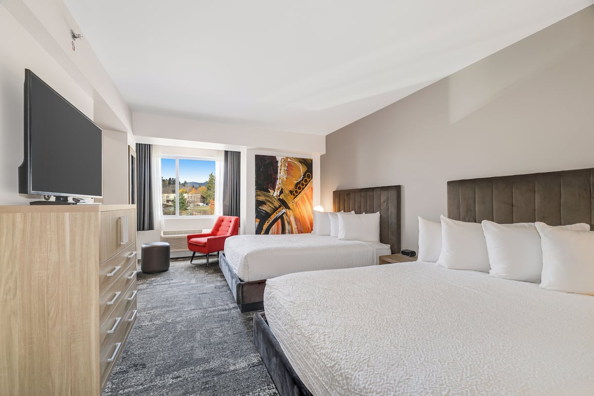 A hotel room with two beds, a TV, a red chair, and a large window with a view of the outdoors.
