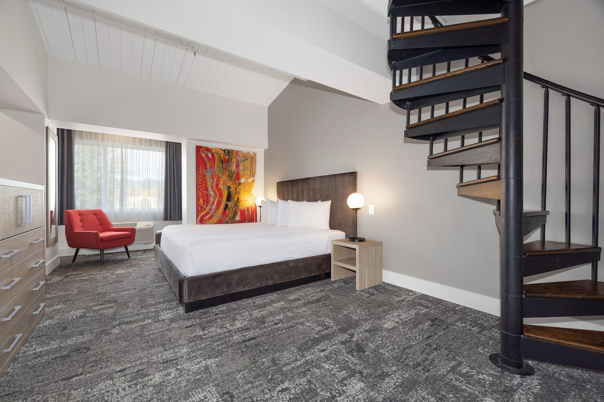 A modern hotel room with a large bed, a red armchair, a wooden desk, and a staircase.