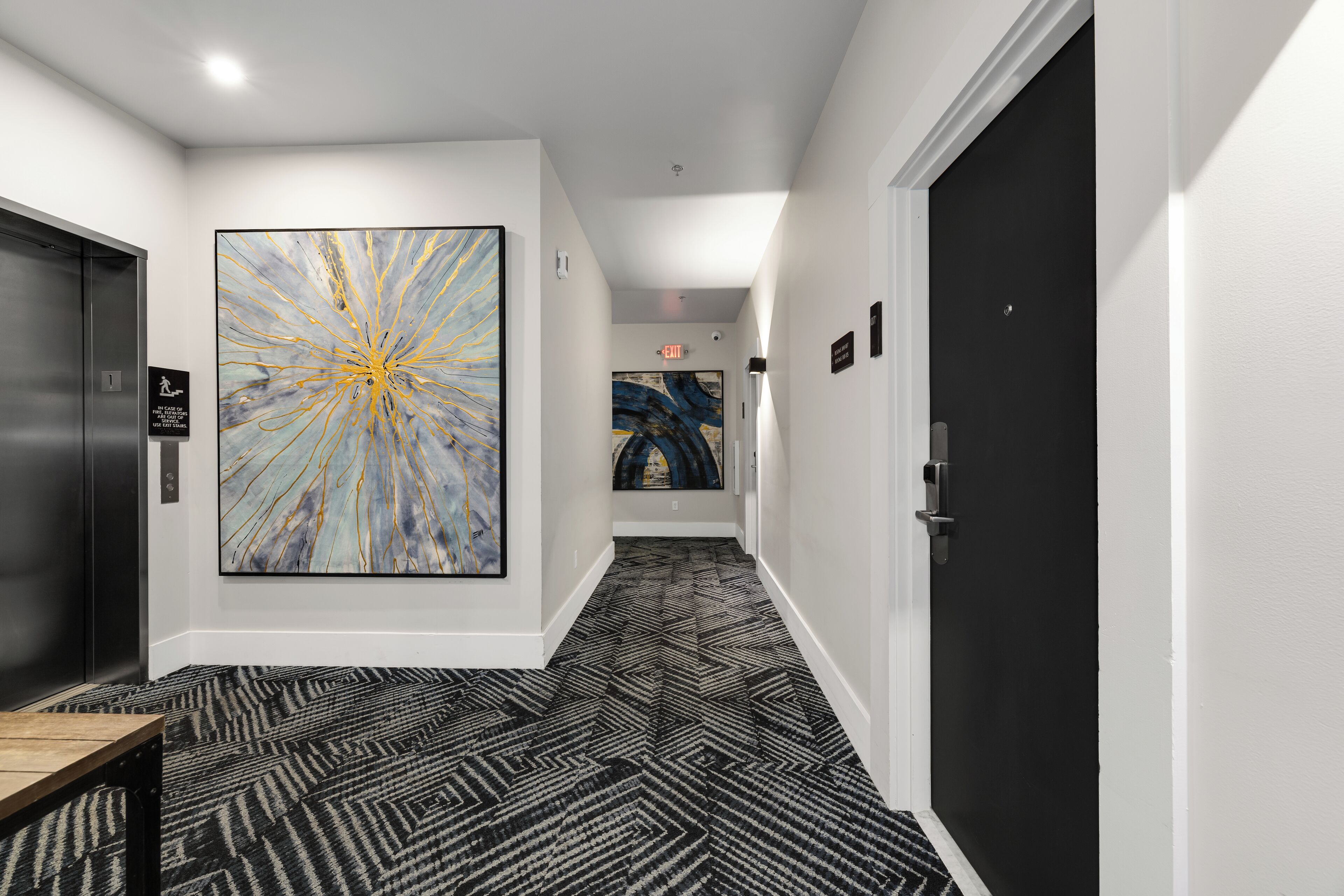 A hallway with a black and white patterned carpet, a large abstract painting on the left wall, and a black door on the right.