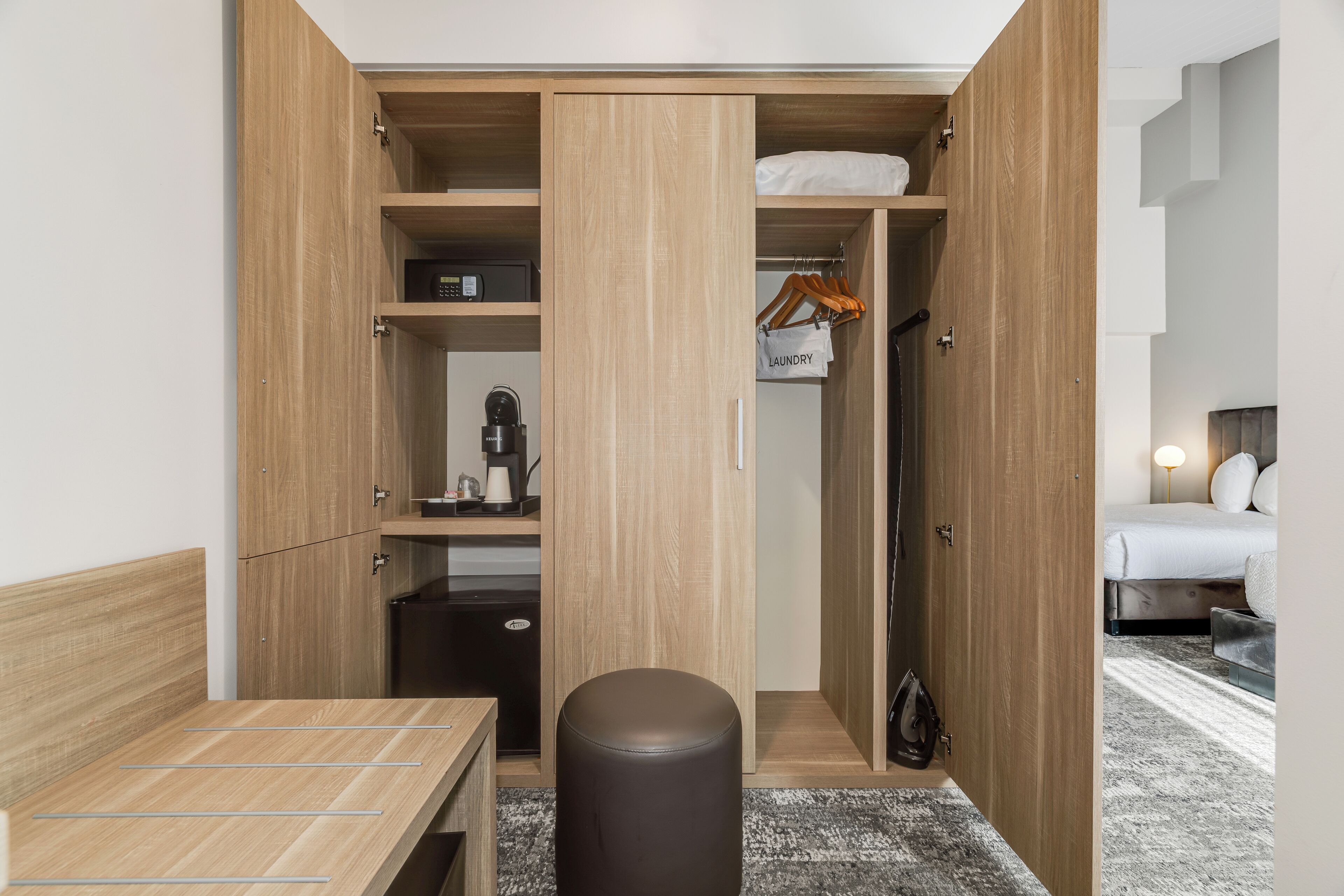 Camera d'albergo compatta con un armadio in legno, un tavolino, un pouf nero e un letto con tappeto grigio.