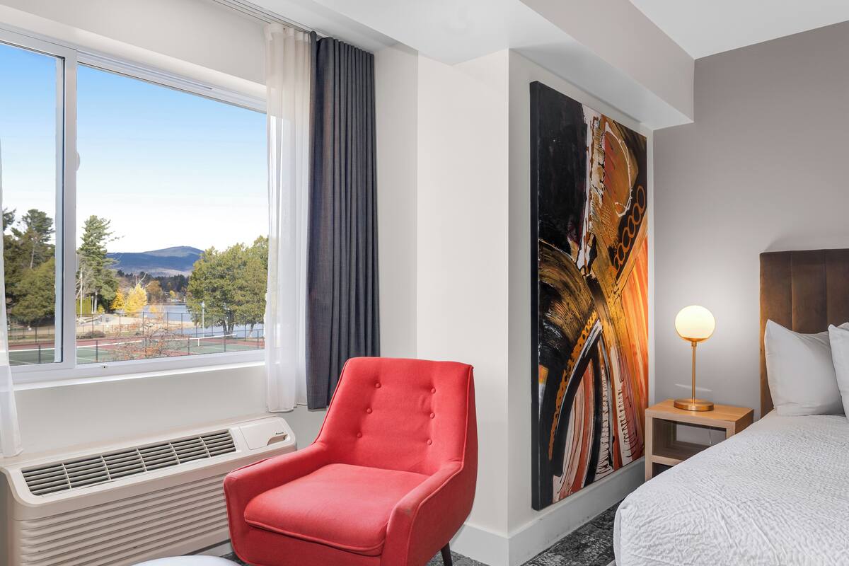 A hotel room with a large window, a red armchair, a bed with white bedding, a nightstand with a lamp, and a large abstract painting on the wall.