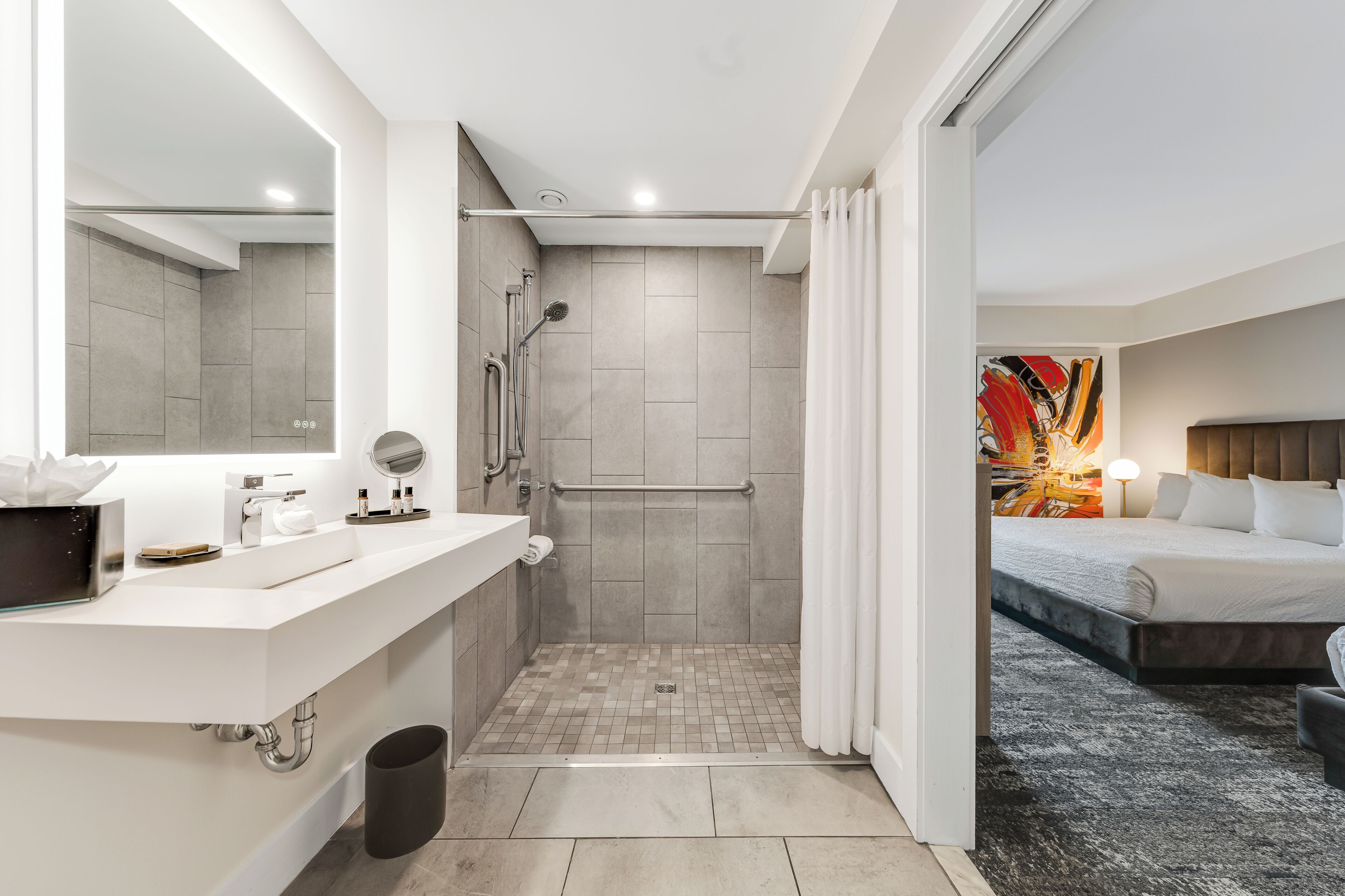 A modern bathroom with a shower, toilet, and sink, and a bedroom with a bed and a painting.
