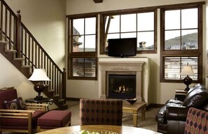 A cozy living room with a fireplace, a television mounted above it, and a staircase leading to an upper level.
