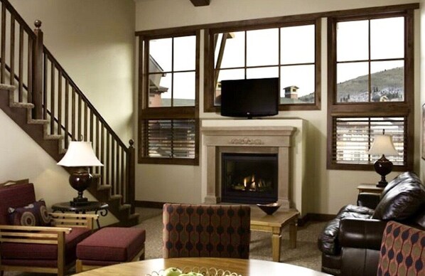 A cozy living room with a fireplace, a television mounted above it, and a staircase leading to an upper level.