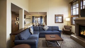 Ein geräumiges Wohnzimmer mit einer großen Eckcouch, einem Couchtisch und einem Kamin. Im Hintergrund ist eine Küche zu sehen.