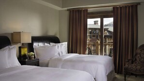 Ein Hotelzimmer mit zwei Betten, einem Nachttisch mit Lampe und Blick auf ein Gebäude durch ein mit Vorhängen versehenes Fenster.