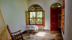 A room with a wooden chair, a small table with a floral tablecloth, a window with wooden shutters, and a red door. - Matagalpa la Tierra Land Of Dreams (San Ramon)