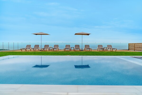 A swimming pool with lounge chairs and umbrellas, surrounded by a wooden fence and a clear view of the sea.