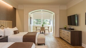 Habitación de hotel con dos camas, zona de comedor con mesa y sillas, televisor montado en la pared y un ventanal con vistas a árboles.