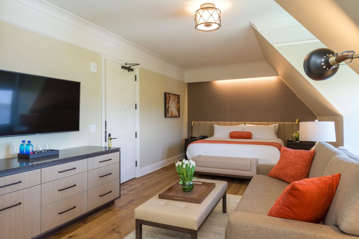 A modern hotel room with a flat-screen TV, a bed with orange pillows, a sofa with orange cushions, a wooden coffee table, and a desk with drawers.