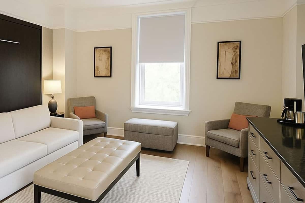 A modern living room with a white sofa, a beige ottoman, two armchairs, a coffee table, and a dark wooden cabinet. There are two framed artworks on the walls, a window with blinds, and a lamp on a side table.
