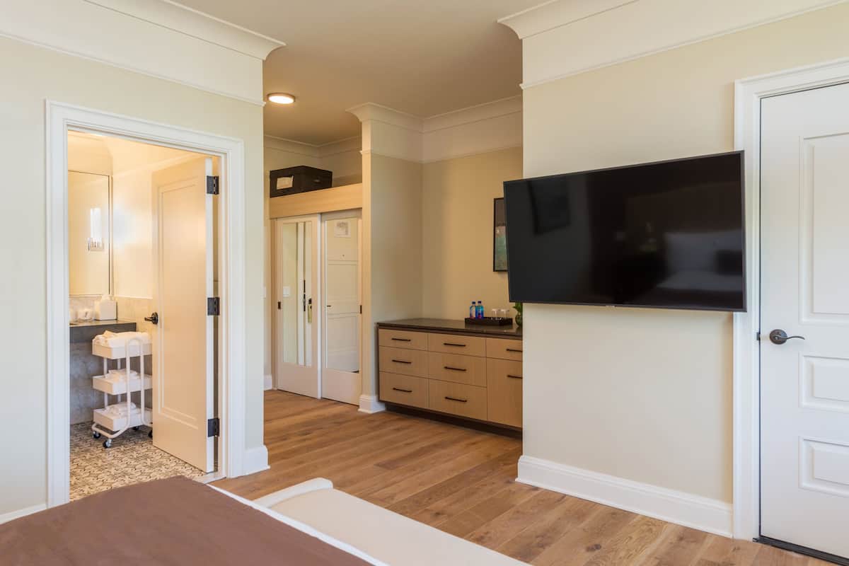 A hotel room with a wooden floor, a flat-screen TV mounted on the wall, a white door, and a small kitchen area with cabinets and a countertop.