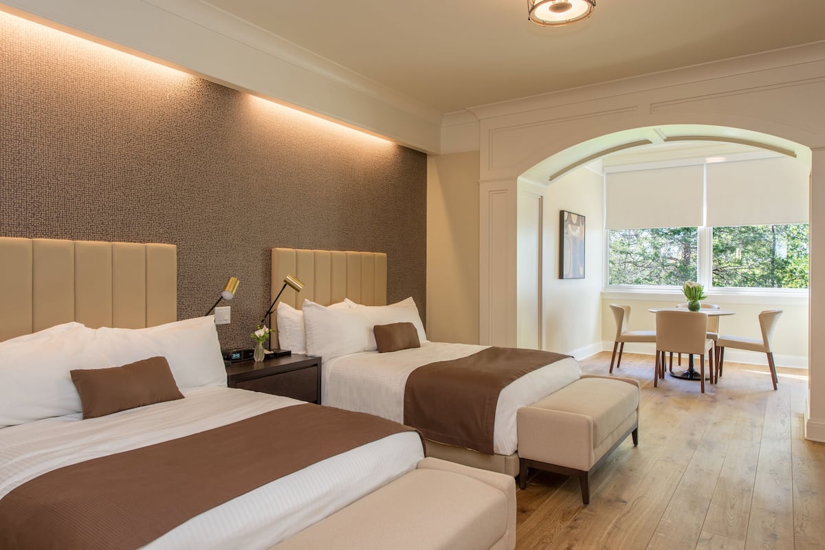 A hotel room with two beds, a sitting area with a table and chairs, and a large window providing a view of the outdoors.