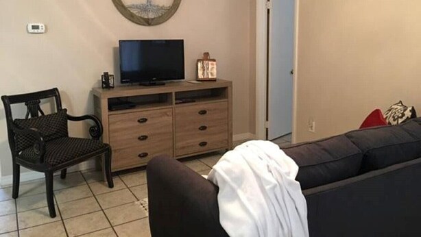 Un salon avec un téléviseur posé sur un meuble en bois, un fauteuil en osier, un canapé foncé et une porte donnant sur une autre pièce.