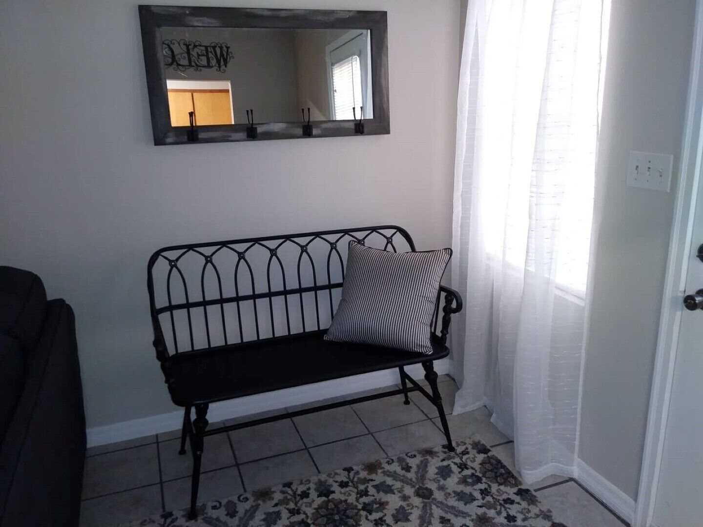 A room with a black metal bench, a patterned rug, a mirror on the wall, and a door with a white frame.