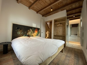 A bedroom with a large bed, wooden ceiling beams, a wooden floor, a painting on the wall, and a walk-in closet with a mirror and shelves. - Award winning luxury, renovated ‘flowerbulb barn’ near beach and Keukenhof (10p) (Noordwijk)