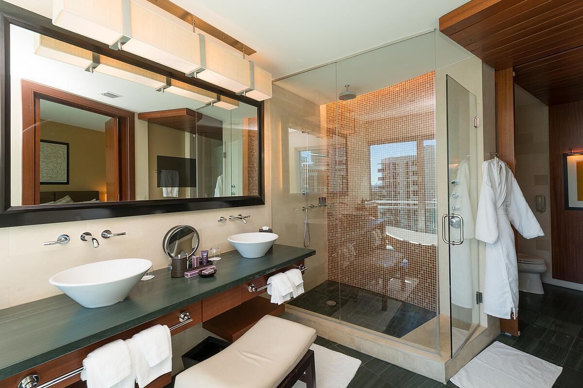Un baño moderno con un espejo grande, dos lavamanos, un banco y una ducha con paredes de vidrio.