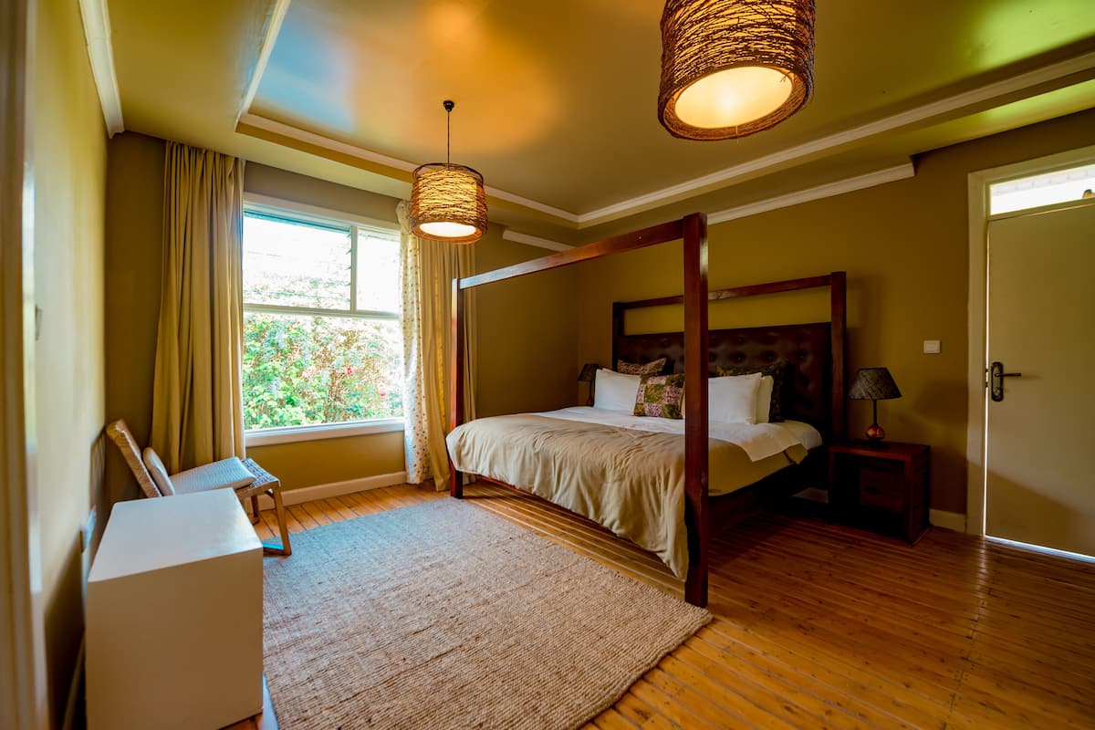 Una cama con dosel, cabecero de madera, mesita de noche con lámpara y vistas al verdor desde la ventana.