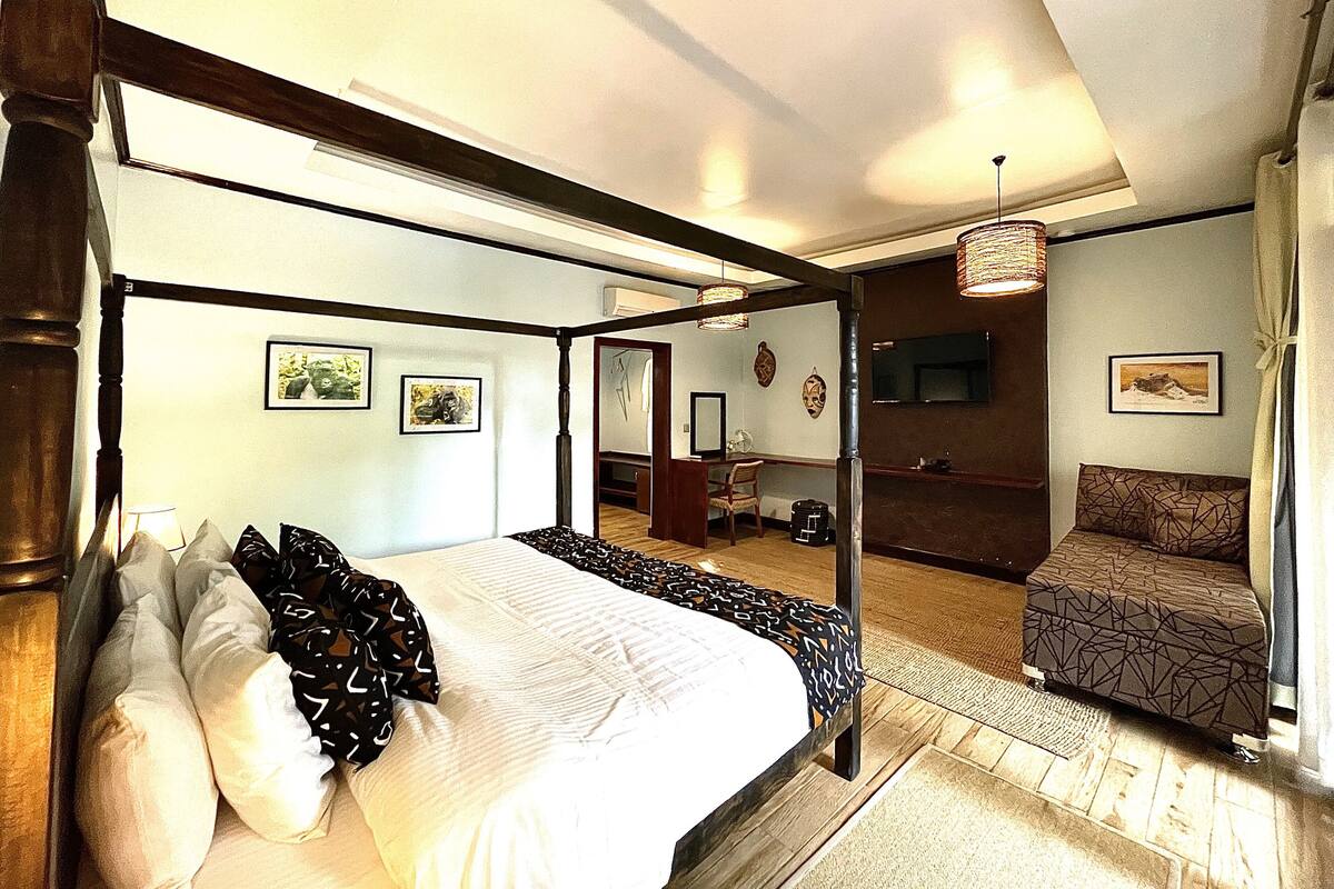 Una cama con dosel, con cubrecama blanco y almohadas estampadas negras, suelo de madera, un sillón estampado y un televisor en la pared.